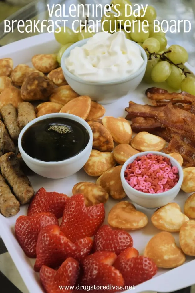 Heart-shaped pancakes, heart-shaped strawberries, sausage, bacon, green grapes, and bowls of sprinkles, maple syrup, and whipped cream on a white board with the words "Valentine's Day Breakfast Charcuterie Board" digitally written on top.