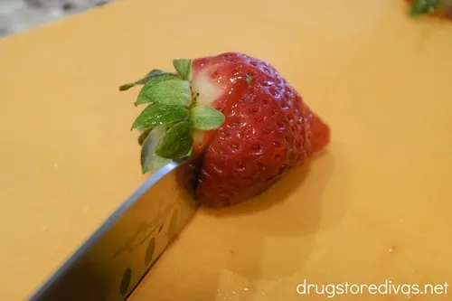 A knife cutting off the top of a strawberry on an orange cutting board.