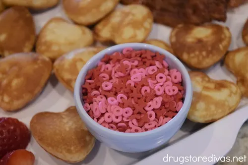 Red and pink X and O sprinkles in a blue bowl with mini heart-shaped pancakes around it on a white serving tray.
