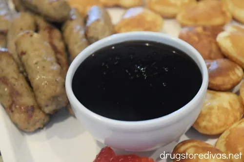 Maple syrup in a blue bowl with mini heart-shaped pancakes and breakfast sausage links around it on a white serving tray.
