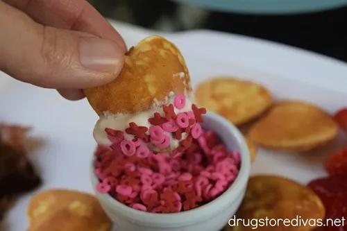 Finger dipping a mini pancake with whipped cream on it into a bowl with pink and red X O sprinkles over a breakfast charcuterie board.
