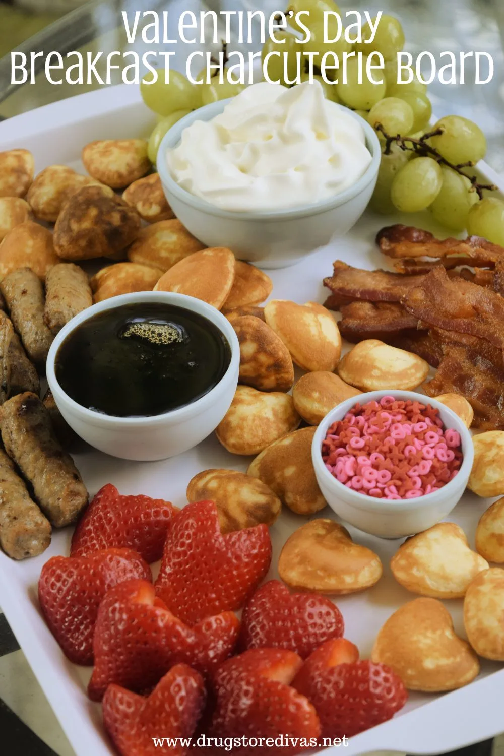 Heart-shaped pancakes, heart-shaped strawberries, sausage, bacon, green grapes, and bowls of sprinkles, maple syrup, and whipped cream on a white board with the words 