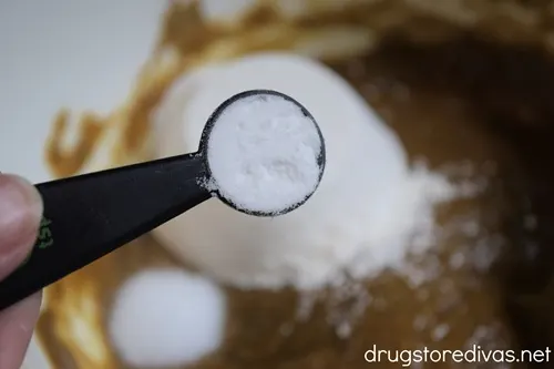 A measuring spoon with a white powder in it above a white bowl with white powder and a brown dough mixture in it.