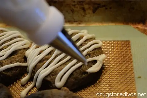A piping bag filled with white chocolate piping white lines onto brown cookies.