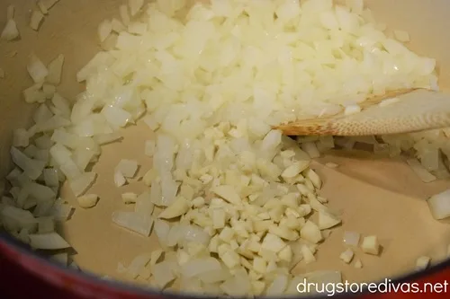Diced onions and garlic in a red Dutch oven being stirred with a wooden spoon.