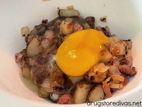 Cooked diced potato, onions, and ham with a raw egg on top in a white bowl.