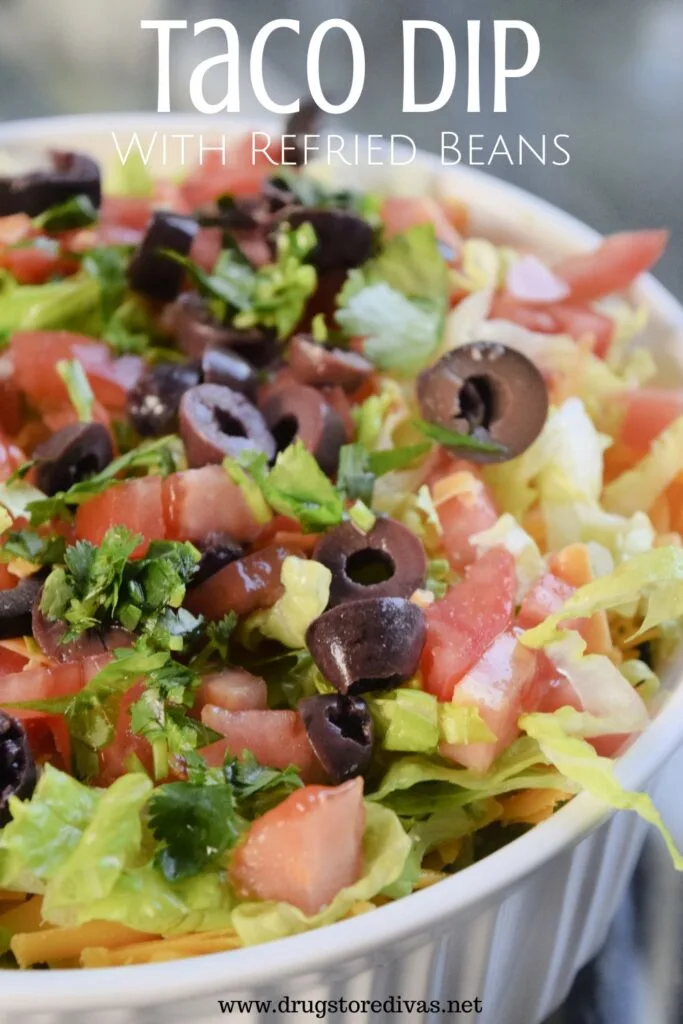 A white, round casserole pan with lettuce, tomatoes, sliced black olives, and cilantro showing and the words "Taco Dip with refried beans" digitally written on top.