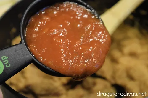A measuring cup filled with red salsa about to be poured into a pot with beans and a wooden spoon it in.