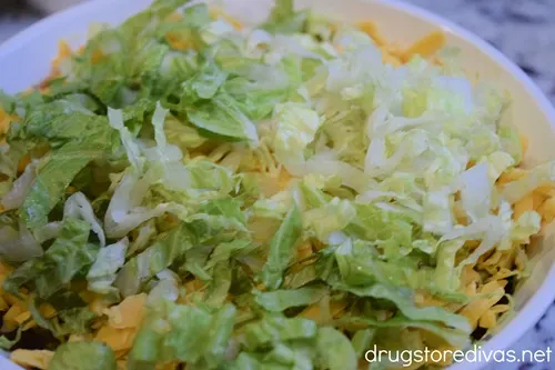 Shredded lettuce on top of shredded cheddar cheese in a white, round casserole pan.