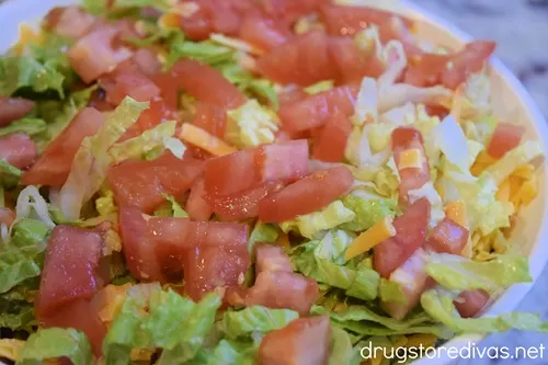 Shredded lettuce and diced tomatoes on top of shredded cheddar cheese in a white, round casserole pan.