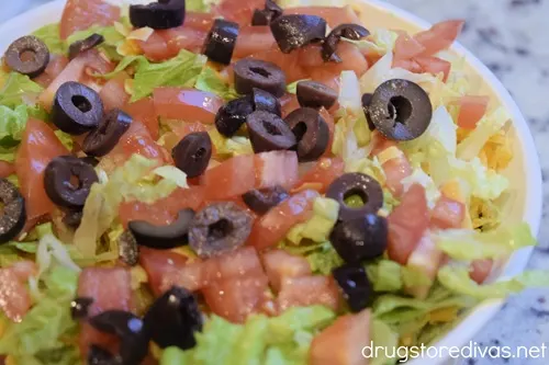 Shredded lettuce, diced tomatoes, and sliced black olives on top of shredded cheddar cheese in a white, round casserole pan.