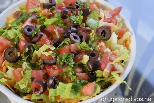 Shredded lettuce, diced tomatoes, sliced black olives, and chopped cilantro on top of shredded cheddar cheese in a white, round casserole pan.