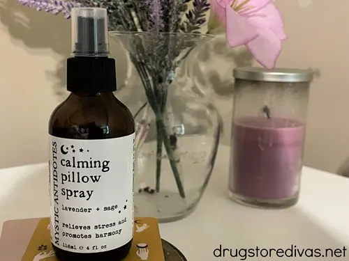 Calming pillow spray on a white table with a flower vase and purple candle behind it.