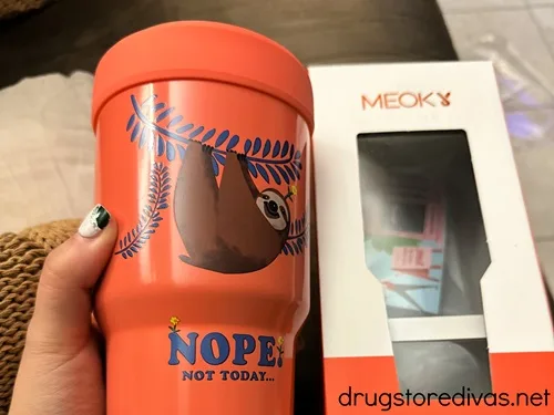 A coral colored coffee cup insulator with a sloth on it that says nope next to the box it came in.