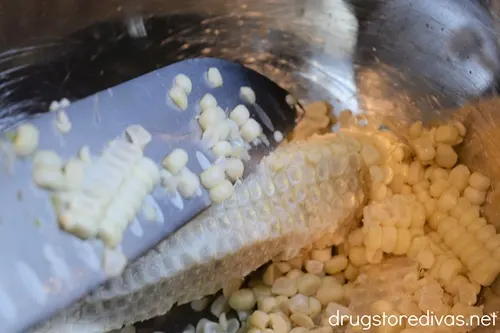 A large knife cutting the corn kernels off an ear of corn and into a bowl.