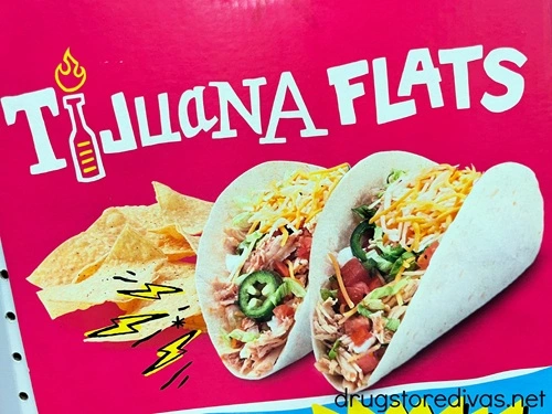A pink sign that says Tijuana Flats with two loaded soft tacos and tortilla chips under it.
