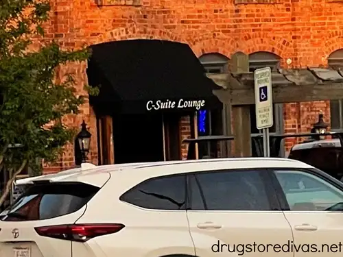 A brick building with an awning on it that says C-Suite Lounge with a white car in a parking lot in front of it.