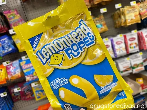 A blue and yellow bag of Lemonhead Ropes candy being held by a hand in front of other candy in the candy aisle of a store.
