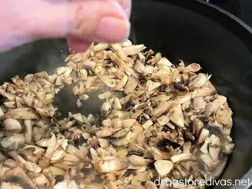 Cooked mushrooms in a pan with a hand sprinkling seasoning over them.