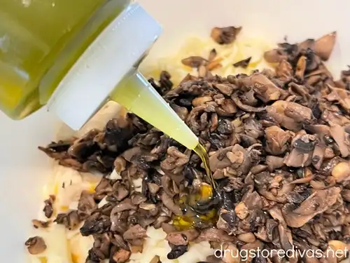 Olive oil from a squirt bottle being poured on cooked mushroom pieces and shredded mozzarella cheese on top of biscuit dough pieces in a white bowl.