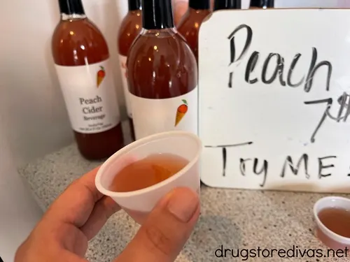 A hand holding a small tasting cup filled with a peach drink with bottles behind it.