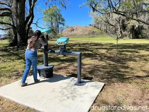 A woman in a gray sweatshirt and blue jeans standing at a viewfinder, looking through at a mound of dirt, at Santee National Wildlife Refuge in Santee, SC.
