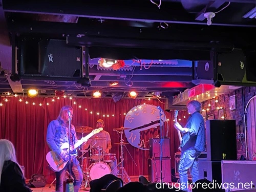 A three piece band on an indoor stage with an emblem that says The Rusty Nail behind them.