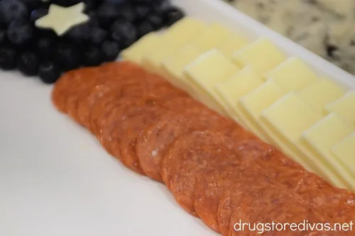 Blueberries with stars on top in a corner and slices of white cheese in two rows, then pepperoni rows, on a white tray.