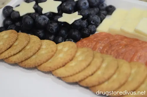Blueberries with stars on top in a corner and slices of white cheese in two rows, then pepperoni rows, then a row of Ritz crackers, on a white tray.