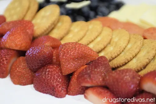 Blueberries with stars on top in a corner and slices of white cheese in two rows, then pepperoni rows, then a row of Ritz crackers, and a row of strawberry halves, on a white tray.