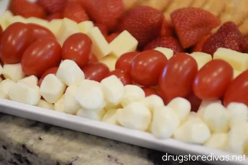 A row of strawberry halves, a row of cubed white cheese, a row of grape tomatoes, and a row of mozzarella pearls on a white tray.