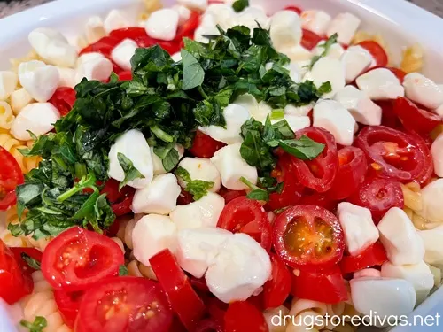 Chopped basil, mozzarella pearls, sliced grape tomatoes, and cooked rotini pasta in a white bowl.