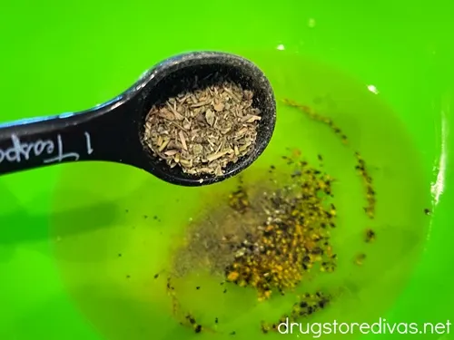 A measuring spoon adding seasoning to a green bowl filled with oil and seasoning.