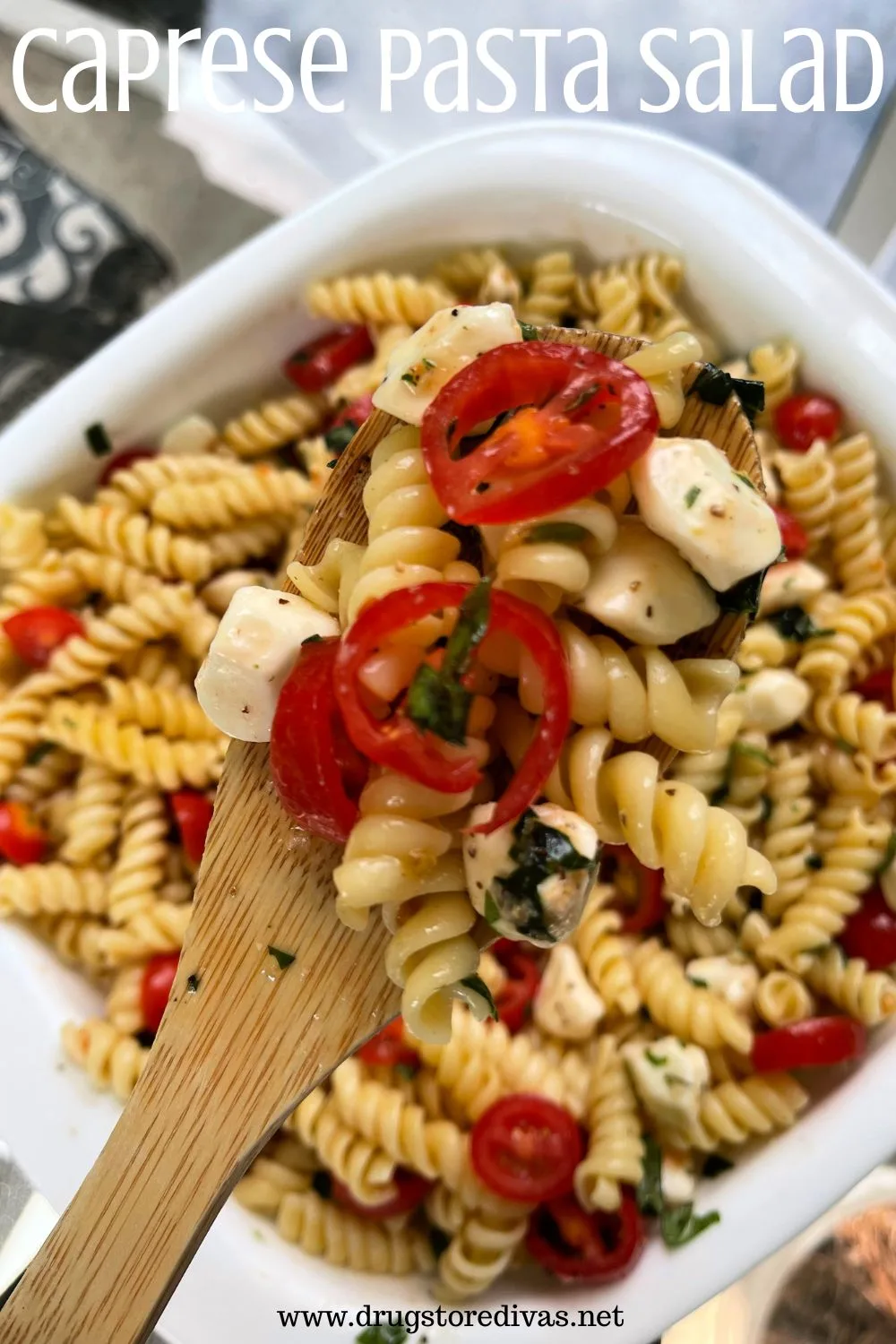 A wooden spoon holding pasta, grape tomato slices, and mozzarella pearls over a bowl containing the same with the words 
