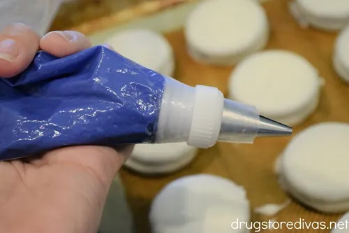 Melted blue candy in a piping bag being held over a bunch of white cookies on a baking mat.