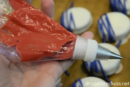 Melted red candy in a piping bag being held over a bunch of white cookies with blue lines on them on a baking mat.