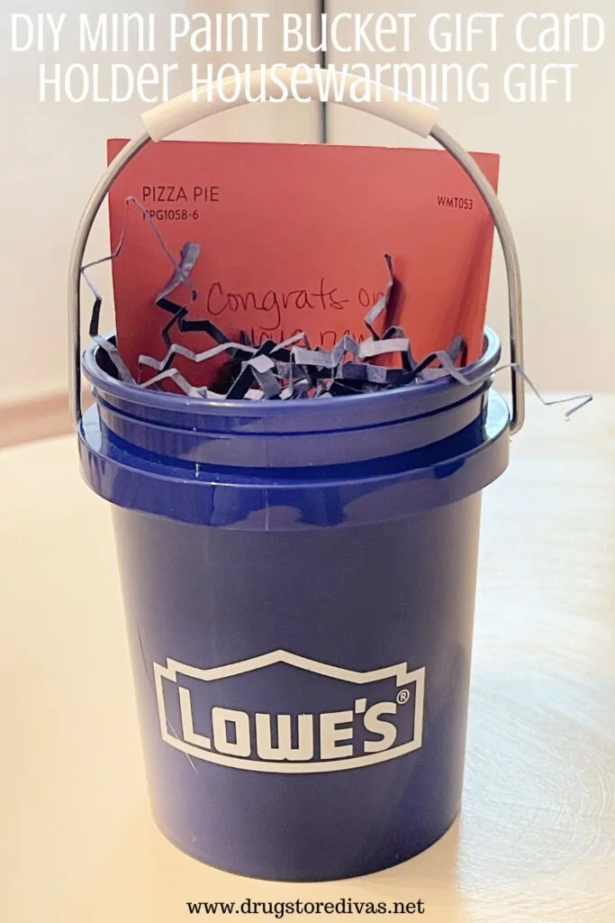 A blue mini paint bucket from Lowe's with a red-ish card in the back and paper shred inside with the words "DIY Mini Paint Bucket Gift Card Housewarming Gift" digitally written above it.