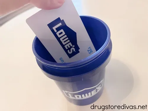 Fingers putting a $25 Lowe's gift card into a blue mini paint bucket from Lowe's.