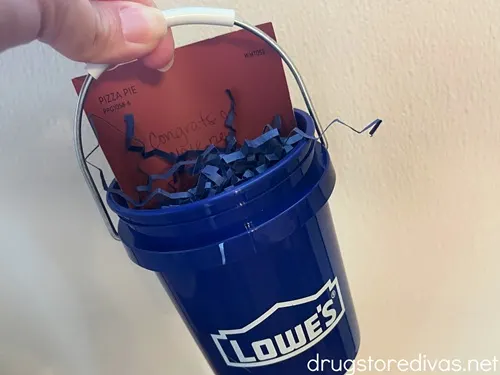 A hand holding a blue mini paint bucket with blue paper shred and a salmon colored piece of paper in it.