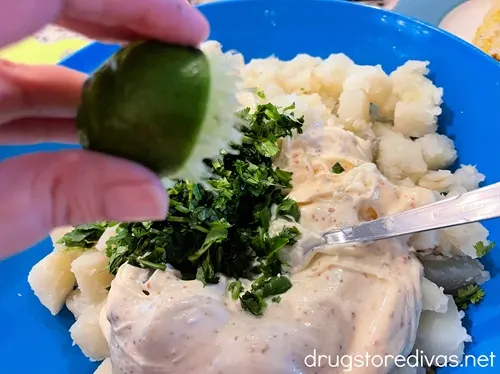 Fingers squeezing half a lime into a blue bowl that's filled with cilantro, a mayonnaise dressing, and cooked potato cubes.