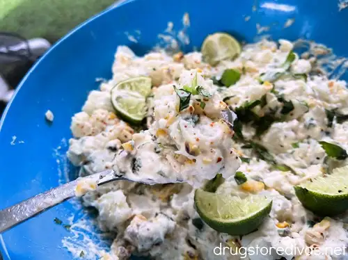 A spoon holding potato salad over a blue bowl filled with Mexican Street Corn Potato Salad, garnished with cilantro, lime wedges, and Tajin seasoning.