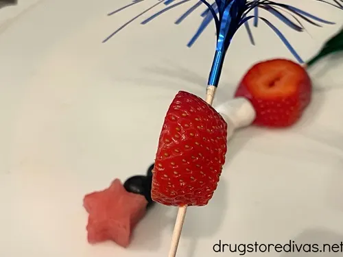 A piece of strawberry on a skewer with blue foil at the top with a fruit skewer behind it.
