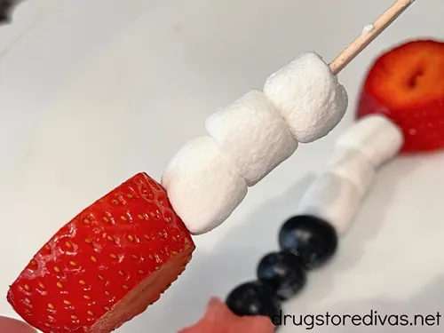 A piece of strawberry and three marshmallows on a skewer with a fruit skewer behind it.