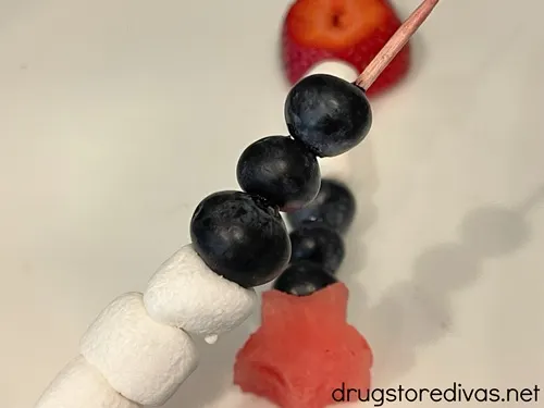 Three mini marshmallows and three blueberries on a skewer with a fruit skewer behind it.