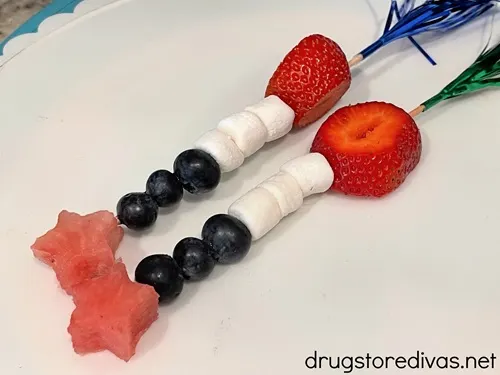 Two fruit skewers, with foil tops, with a slice of strawberry, three marshmallows, three blueberries, and a star-shaped watermelon slice on a white plate.