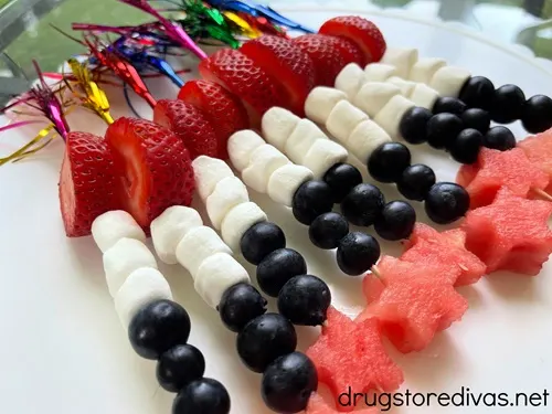 Nine fruit skewers, with foil tops, with a slice of strawberry, three marshmallows, three blueberries, and a star-shaped watermelon slice on a white plate.