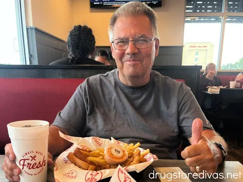 A man in a gray shirt sitting in a booth at Slim Chickens in front of a plate with chicken fingers, fries, and sauce, with a drink in a white cup, giving a thumbs up.
