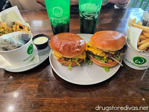 Two cheeseburgers on a plate, with bowls of fries and both sides, and two green soda cups behind them, all branded with Wahlburgers.