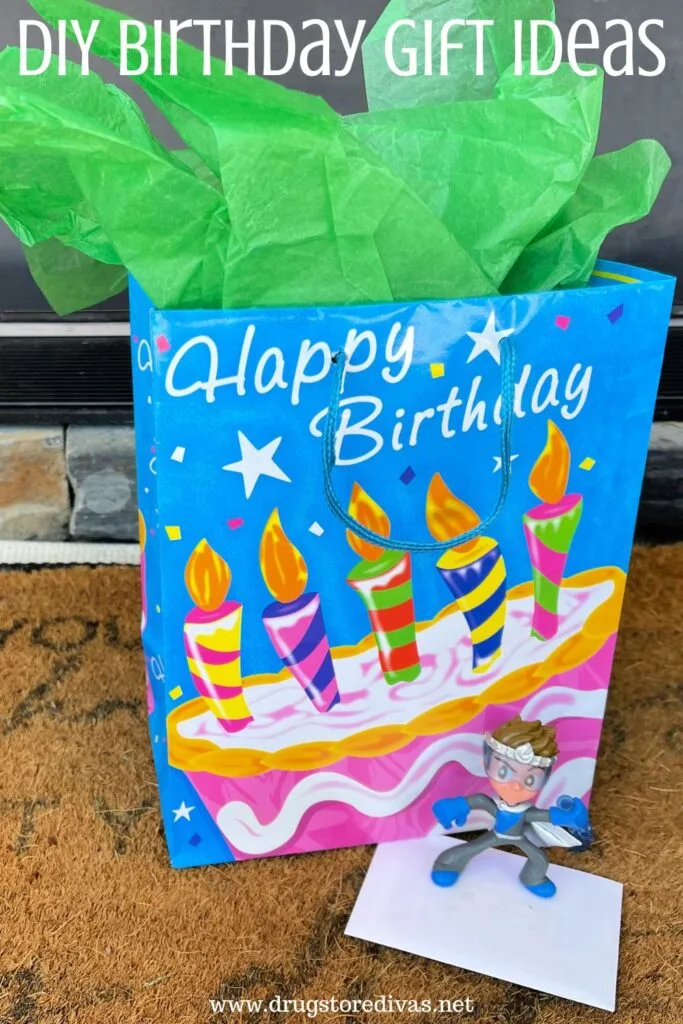 A gift in a Happy Birthday bag with a card and small action figure in front on a doormat with the words "DIY Birthday Gift Ideas" digitally written on top.