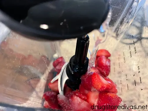 Water in a black measuring cup being added to a blender that has strawberries in it.